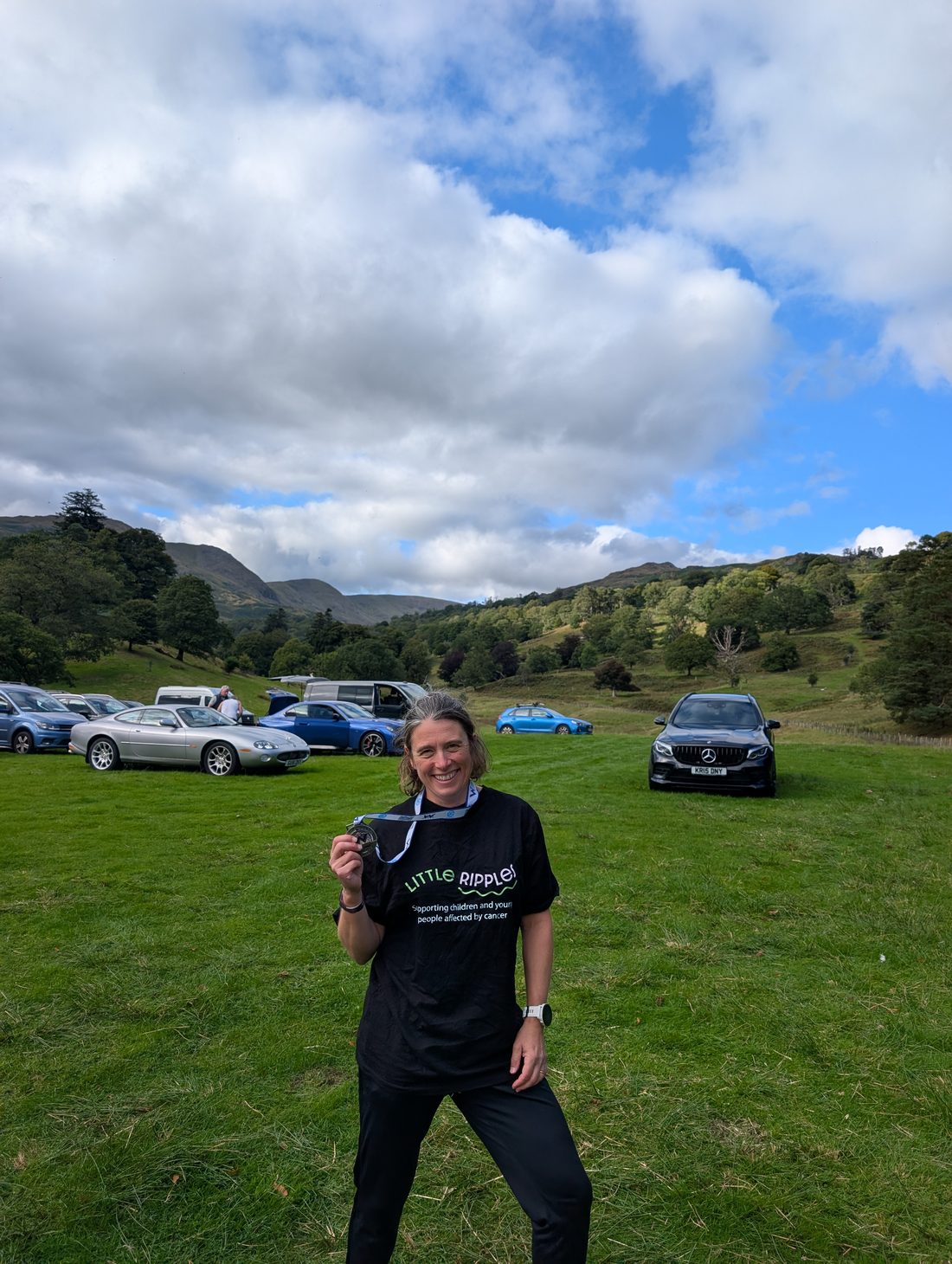 A Little Ripples fundraiser in a branded shirt holding up her finisher medal after a run in the Lake District
